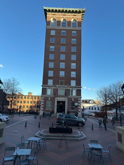A tall, brick building dominates the scene, its height emphasized by the upward angle of the shot. A clock is visible on the facade. Below, a brick-paved street features a fountain or monument in the foreground. Sidewalk tables and chairs are arranged nearby. Several people are walking on the street, and a dark SUV is parked near the building's entrance. The sky is clear blue. Another building is visible across the street. Bare tree branches frame the scene.