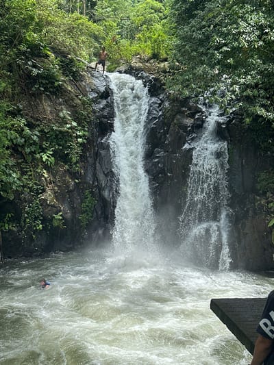 This image depicts a multi-tiered waterfall in a lush, tropical environment, identified as Ambengan, Indonesia. A shirtless male, wearing dark shorts, stands atop the dark, jagged rocks at the precipice of the main waterfall, looking downwards. A powerful stream of white, turbulent water plunges from this point into a roiling pool below. To the right, a secondary, smaller cascade also flows down the dark rock face. In the turbulent water of the pool at the base of the waterfall, an individual is partially submerged, wearing a blue or dark flotation vest, with their head and upper torso visible. In the bottom right foreground, the right shoulder and arm of a third person are visible, wearing a dark t-shirt with white lettering. This individual is positioned on a dark, wet wooden or concrete platform extending into the water. The surrounding environment consists of dense green foliage and trees covering the steep rock formations.