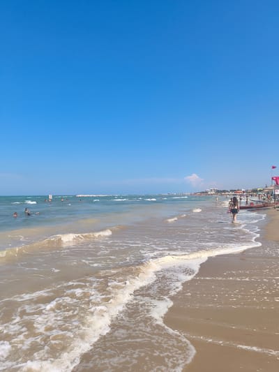 The image depicts a beach scene on a sunny day with a clear blue sky. In the foreground, gentle waves with white foam are washing ashore onto a sandy beach. A woman in a black bikini is walking away from the camera along the water's edge, carrying what appears to be a surfboard or a similar flat object.
Further out in the water, several people are swimming and enjoying the sea. In the distance, a long line of beach umbrellas and buildings suggests a developed coastal resort area. A red flag, possibly indicating a lifeguard station or a warning, is visible on the right side of the image, attached to a structure with the word "SALVATAGGIO" (meaning rescue) on it, further identifying the location as a supervised beach. The water is a light brown color, indicating sand or sediment stirred up by the waves. The overall impression is a typical summer day at a crowded beach resort.