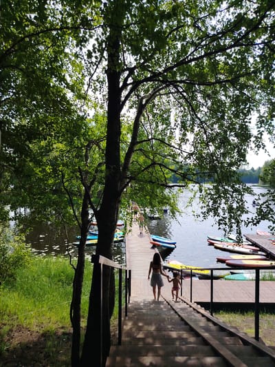In Выльтыдор, Russia, a wooden boardwalk extends into a calm body of water. On the boardwalk, several paddleboards are neatly arranged. Two individuals, a woman and a young child, are walking down a wooden staircase leading from the boardwalk. The scene is bordered by lush green grass and foliage, with trees casting shadows. The water is populated with a few other people engaged in water activities.