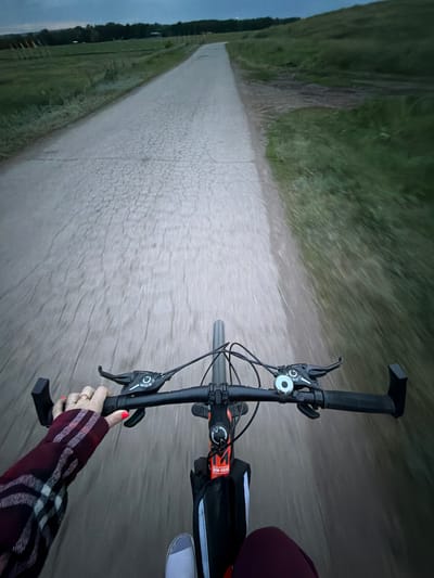 A first-person perspective video shows a person riding a bicycle down a dirt path. The rider's hand, with red nail polish and rings, is visible gripping the handlebars. They are wearing a red and black plaid long-sleeved shirt. The handlebars are black, with brake levers and grips. A black bag is attached to the bicycle frame. The path is surrounded by dry grass and vegetation, with a cloudy sky visible in the background. The lighting suggests it is either early morning or late evening. The scene appears to be in a rural or natural setting. There is a visible text on the bag attached to the bike that reads "HW-560".