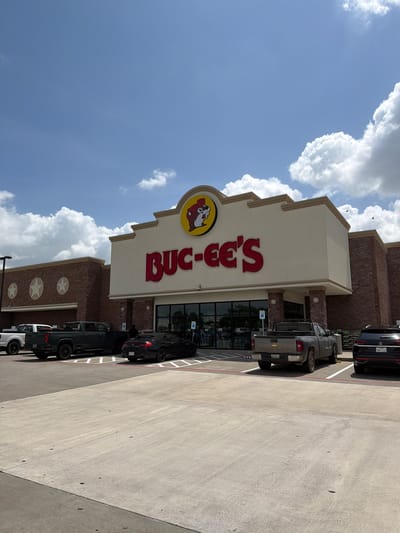 A vertical, low-angle shot captures the exterior of a Buc-ee's convenience store. The iconic Buc-ee's beaver logo, a cartoon beaver wearing a red bow tie, is prominently displayed on a yellow circular sign. Red, block letters spelling "Buc-ee's" are mounted above the store's entrance. The building features brown brick accents and a light-colored facade. Several vehicles, including a black SUV and two dark-colored pickup trucks, are parked in the lot. A light-colored pickup truck is partially visible at the bottom of the frame. The scene is set against a bright blue sky dotted with fluffy white clouds, suggesting a sunny day. The concrete ground of the parking lot is visible in the foreground and to the right. The overall atmosphere appears to be that of a typical daytime scene at a popular roadside stop. The image is located in Katy, United States of America.