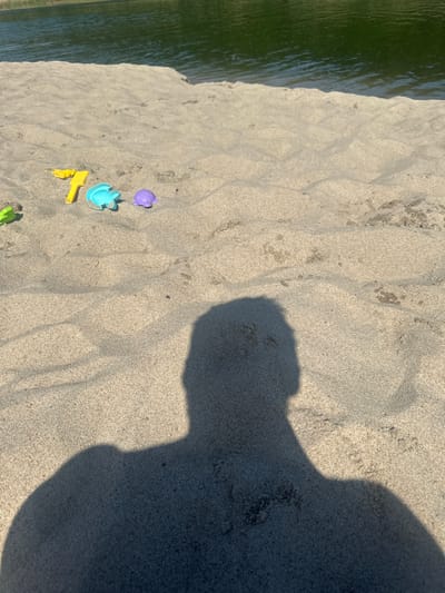 The image shows a close-up view of sand with several colorful plastic beach toys, including a yellow shovel, a blue turtle mold, and a purple turtle toy. In the background, a body of dark green water, likely a lake or river, meets the sandy shore. The shadow of a person is prominently cast across the sand in the foreground, suggesting the photograph was taken on a sunny day. The scene appears to be a recreational area, possibly a beach within the city of Yantarny, Russia, known for its sandy shores and proximity to water bodies.