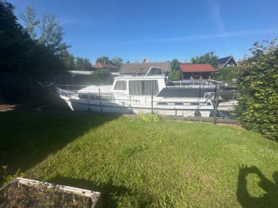 This image depicts a residential area in Ossenzijl, Netherlands, on a sunny day with a clear blue sky. In the foreground, a well-maintained lawn is visible, with a planter box containing purple flowers. A shadow of a person's hand, appearing to form bunny ears, is cast upon the grass.
The main subject of the image is a white houseboat, identified by the name "Perdix", docked behind a metal fence. The boat has several windows with drawn curtains and a dark-colored hull. Beyond the boat, a row of houses with red and brown tiled roofs are nestled amongst lush green trees. A fence separates the property with the boat from the neighboring houses. The overall impression is one of a peaceful, suburban environment.