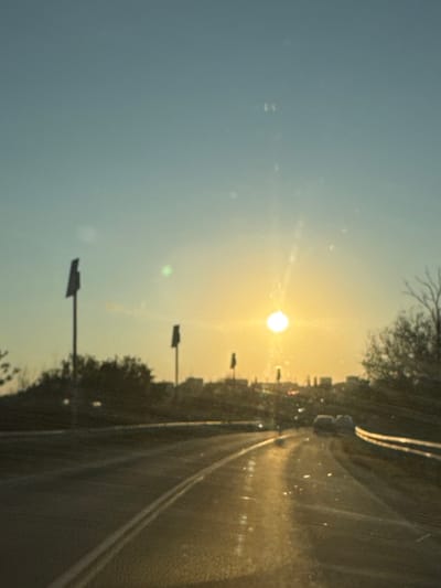 The image depicts a road in the city of Ol'ginskaya, Russia, at sunset. The sun, a bright orb, is low on the horizon, casting a warm, golden glow across the scene. The sky is a clear, pale blue, transitioning to yellow near the sun. The road, which appears to be paved, curves gently to the right. The wet surface of the road reflects the sunlight and the surrounding environment, creating streaks of light.
On either side of the road, there are dark shapes of trees and vegetation. In the background, a city skyline is visible, with buildings silhouetted against the bright sky. There are also two tall, slender poles with rectangular signs at the top, positioned along the left side of the road. These may be road signs or traffic signals.
In the distance, several cars are visible on the road. Their headlights are illuminated, suggesting that it is either dusk or a dim setting. The overall impression is one of a peaceful, late afternoon or early evening drive through a semi-urban landscape. The image is slightly out of focus, giving it a dreamlike quality.