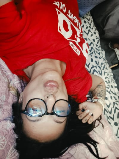 The image depicts a woman lying down on a bed. She is wearing a red t-shirt with white lettering that appears to say "NIGHT OFF SMIRNOFF." She has black-rimmed glasses, a nose ring, and dark hair. Her nails are painted with a black and white polka dot pattern. The bed has patterned sheets. There are tattoos visible on her arm. The overall scene appears to be indoors, possibly a bedroom. There is no information within the image to suggest a location in Bridlington, United Kingdom.