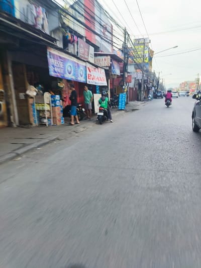 The image shows a street scene in Bojongsoang, Indonesia, located at latitude -6.98 and longitude 107.634. There are three people standing on the sidewalk in front of a storefront. Two of the people are men, and one is a woman. The men are both wearing casual clothing, and the woman is wearing a long-sleeved shirt. The storefront has several signs, including one that says "ESE CHANGE". There are several motorcycles parked on the sidewalk, and a few cars are driving down the street. The street is paved with asphalt, and there are power lines overhead. The sky is overcast, and there are no visible trees. The image appears to have been captured from a moving vehicle.