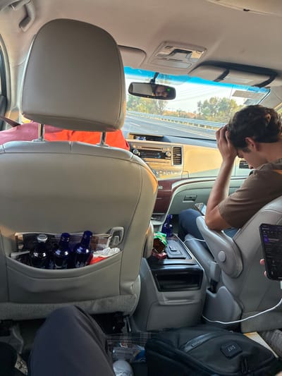 The image depicts the interior of a moving vehicle, identified as being in the vicinity of Santa Ana Xalmimilulco, Mexico. The perspective is from a rear passenger seat looking forward.

In the foreground, the back of a light-colored headrest from the seat directly ahead is visible. The seat-back pocket contains three dark blue bottles, one identified as "Skyy," along with crumpled white material. Below, the legs of a passenger in dark pants and white shoes are partially visible.

To the right, a male passenger in the front seat, wearing a brown shirt and dark pants, is observed with his head down, hands clasped over his head or forehead, suggesting a state of rest, discomfort, or contemplation. Adjacent to him, on the center console, are several snack bags and another dark blue bottle. A white charging cable extends from the console towards the rear right. On the floor to the bottom right, a black bag with "SNF" initials and a plaid object are present. A hand holding a black smartphone displaying a purple-themed music application is partially visible in the lower right.

The vehicle's dashboard and infotainment system are visible between the front seats. The rearview mirror reflects the face of the male driver and displays a green digital "95," indicating speed or a road marker. Outside the windshield, a multi-lane highway with concrete barriers and roadside vegetation is visible under daylight conditions, possibly late afternoon.