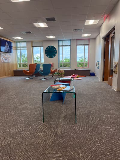The image shows an interior view of what appears to be a waiting area or a children's play area. In the foreground, a low, glass-topped coffee table with a blue base holds several children's books. Beyond this, the room features a patterned carpet, several colorful, modern chairs, large windows with views of greenery, and a large, teal-colored clock on the wall. To the left, a flat-screen television displays a cartoon image. In the background, there are cushioned benches along the wall beneath the windows and some soft play mats on the floor. The overall atmosphere is bright and inviting, suggesting a public space designed for families. The specific location is the City of Chesapeake, United States of America.