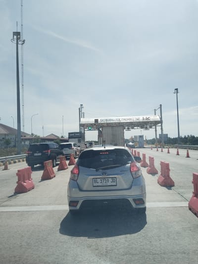 The image shows a line of cars at the Padang Hilir toll gate in Mukee Gogo, Indonesia. A silver Toyota Yaris is in the foreground. Orange traffic barriers are placed along the road. In the background, a gantry with "GERBANG TOL PADANG HILIR" (Padang Hilir Toll Gate) is visible. A tall metal tower stands to the left. The sky is partly cloudy.
