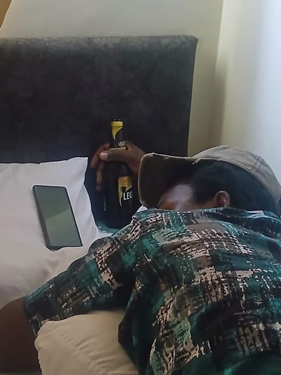 The scene depicts a man wearing a patterned shirt and a cap, holding a bottle of "Leg" beer. A smartphone lies on a white surface. The background includes a dark-colored wall. The city of Heipang, Nigeria, is not discernible from the image.