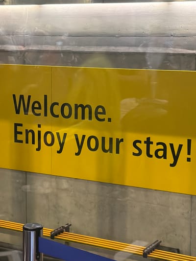 The image shows a yellow sign reading "Welcome. Enjoy your stay!" in black lettering. The sign is located in Kloten, Switzerland, evidenced by the airport code visible in reflections. To the right of the sign is a retractable belt barrier system with yellow and blue belts. The sign is mounted on a grey concrete wall. The scene appears to be inside an airport terminal. No people are directly visible in the image.