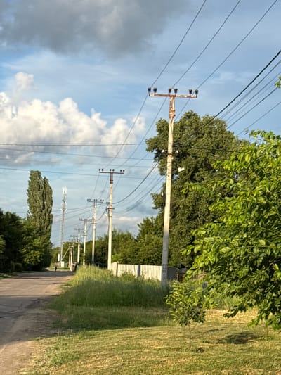 The image depicts a street in Rossosh, Russia, lined with power lines supported by numerous poles. A tall, slender tree stands on the left side of the road, with a cell tower visible further down. The road appears to be asphalt, with some wear and tear. On the right side of the road, there is a large tree and a smaller sapling, with a white fence and tall grass in the background. The sky is partly cloudy, with a mix of blue sky and white clouds. A lone figure is walking down the road in the distance. The scene appears to be an outdoor, rural or suburban setting.