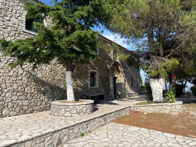 The image depicts the exterior of a stone building, likely a church, in a sunny outdoor setting. The building features a rough-hewn stone facade and a tiled roof. An arched entrance with a mosaic above it is visible on the right side of the building. Trees with lush green foliage frame the building, casting shadows on the stone walls. The ground in front of the building is paved with irregular flagstones, and there is a low stone wall bordering a path. A circular stone planter with a white-painted tree is in the foreground. Another white-painted tree stands to the right, adorned with pink flowers. The overall scene suggests a peaceful and historic location, likely in Greece given the provided location context. The time of day appears to be daytime, with clear blue skies and bright sunlight. There is no visible text in the image. The atmosphere conveyed is one of tranquility and natural beauty.