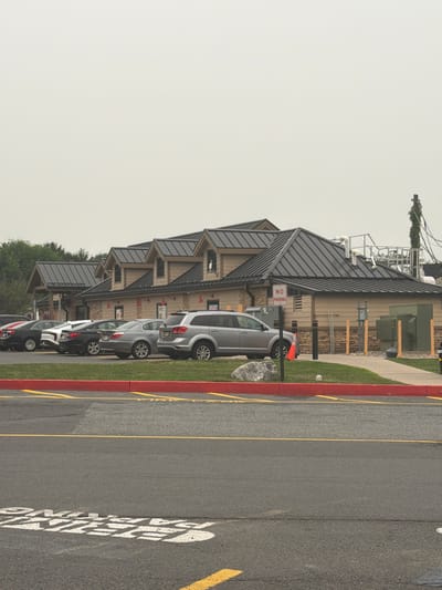 The image shows a parking lot in front of a building with a dark metal roof. Several cars are parked in the lot. A "NO PARKING" sign is visible on a pole. The building appears to be a commercial establishment. The sky is overcast.