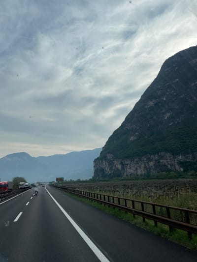 The image depicts a multi-lane highway with several vehicles moving in the same direction. To the left of the highway is a large mountain range. A barrier separates the road from a grassy area with a vineyard. The sky is overcast with clouds. The location is near Mezzocorona, Italy, based on the landscape features.