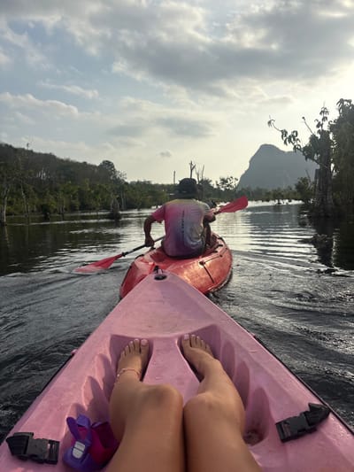 The scene depicts two kayaks on a calm, dark waterway in Ban Khao Klom, Thailand, observed from a first-person perspective within a pink kayak. In the immediate foreground, the bare feet and lower legs of a person are visible, resting inside the pink kayak. The toenails are painted light pink, and a light-colored beaded ankle bracelet is on the right ankle. A purple and pink water shoe rests near the left foot, alongside a black buckle mechanism attached to the kayak's interior.
Directly in front, a red kayak is moving away from the viewer, creating a visible wake. A person is seated in the red kayak, facing forward, and actively paddling with a red double-bladed paddle. This individual wears a dark brimmed hat and a tie-dye t-shirt displaying colors of pink, purple, and white, with partial text "HOLIDAY RESOR" visible on the back.
The waterway is flanked on both sides by dense green foliage, appearing as either lush riverbank vegetation or mangrove trees, with some bare or dead tree trunks protruding from the water. In the distant background to the right, a prominent, steep, karst-like mountain formation rises against an overcast sky. The sky features scattered grey and white clouds, with a brighter area on the right suggesting the position of the sun. The two kayaks are traveling sequentially down the waterway.