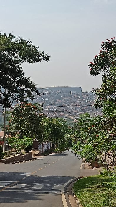 The image displays an urban landscape in Kigali City, Rwanda, captured from an elevated position looking downhill. In the foreground, a paved asphalt road features a double yellow center line, a white dashed shoulder line on the right, and a pedestrian crosswalk marked by white stripes in the lower left. The road is flanked on the right by a concrete curb and a grassy verge, and on the left by a low stone wall with a subsequent vertical wall adorned with multiple posters. Lush green trees, some displaying red flowers, line both sides of the road, casting shadows.
The midground reveals a dense residential and commercial area comprising numerous buildings on undulating hills, interspersed with vegetation. In the distant background, a prominent grey-colored stadium with a flat roof, identifiable as the BK Arena (formerly Kigali Arena), dominates the skyline atop a high hill. The sky above is light grey and appears hazy. No discernible individuals or vehicles are present in the visible portion of the scene.