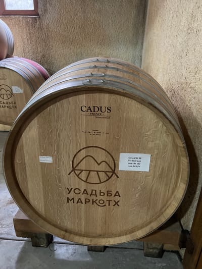 The image displays the head of a large, light-colored oak wine barrel, centrally positioned and occupying the majority of the frame, within a winery located in Vinogradnoe, Russia. The barrel, secured by several polished metal hoops with visible rivets, rests on two dark wooden chocks on a grey concrete floor.
Engraved on the barrel head are the words "CADUS FRANCE Sensoriel" at the top, followed by smaller text indicating "Equilibre French Oak 30 months air dried M Pur Process." Below this is a circular logo depicting stylized mountains with a path, and beneath the logo, the Cyrillic text "УСАДЬБА МАРКОТХ" (Usadba Markotkh).
Two rectangular white labels are affixed to the barrel head. The left label contains handwritten Cyrillic: "Вино 2021" (Wine 2021) and "залито 13.09.23." (filled 13.09.23). The right label is printed with Cyrillic text: "Бочка № 36" (Barrel No 36), "V = 50.0 дал" (Volume = 50.0 decaliters), "инв. № 162" (Inv. No 162), and "зав. № б/н" (Factory No b/n).
In the background, other barrels are partially visible, including one with a red-painted hoop section. The walls are rough-textured, appearing to be light brown or beige stucco. A small, rectangular window with a brown frame is visible in the upper left background, suggesting natural light entering the space. No individuals are present in the scene.
