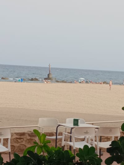 The image depicts a beachfront scene in Águilas, Spain. In the foreground, a few white plastic chairs and tables are arranged on a patio, with lush green foliage obscuring parts of the lower frame. Beyond the patio, a sandy beach stretches towards the sea. Several beach chairs are visible on the sand, some occupied by people sunbathing. A small lighthouse-like structure stands in the water, surrounded by rocks and the sea. The sky is overcast, and the water appears calm. In the distance, more people are scattered along the beach.