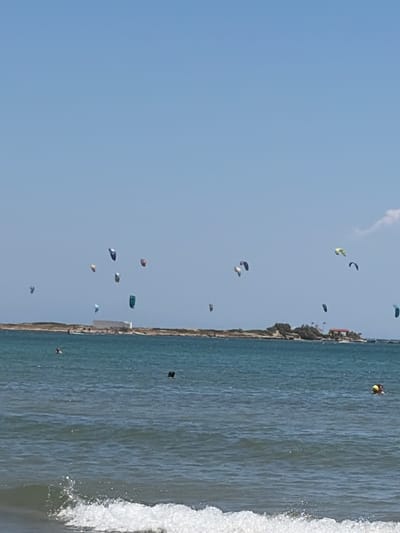 In the city of Artemida Municipal Unit, Greece, a bright, clear sky dominates the upper portion of the image. The sea is a deep blue, with visible waves and whitecaps near the foreground, indicating a breezy day. A distant, sandy landmass stretches across the middle ground, dotted with sparse vegetation and some buildings, including a white structure and a reddish-roofed building. The primary activity depicted is kitesurfing, with numerous colorful kites of various sizes and shapes scattered across the sky above the water and the distant shore. Some kites are closer to the water, while others are much higher. In the sea, several individuals are present. Two figures are visible in the water, one appearing to be swimming, and another further out with a yellow object, possibly a flotation device or ball. A small, dark object, possibly a boat or a person, is also visible in the water closer to the shore.