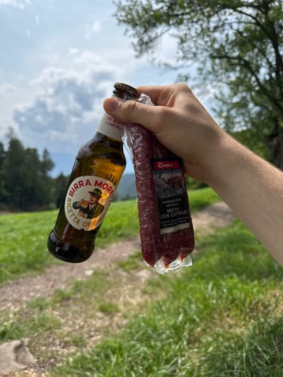 The image shows a person's hand holding a package of "Rinn's Kaminwurzen Affilfülle mit Fisch con Camiolo" sausage and a bottle of "Birra Moretti" beer. The bottle is brown glass with a white label and a gold cap. The sausage is red and has a white label with black text. Both items are being held by a person's hand. The scene appears to be a hiking trail in Costalovara, Italy. There is green grass and trees in the background. The sky is blue and cloudy. The location is coordinates 46.525, 11.424.