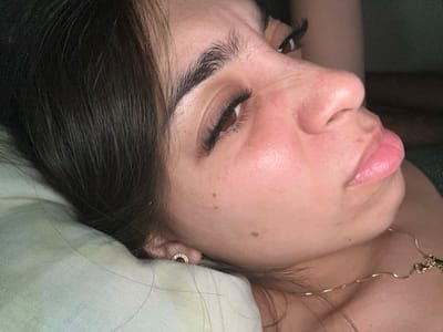 A close-up side-profile shot of a woman lying on a pillow, with dark hair near her face. She has long eyelashes, full lips, and a gold chain around her neck. Her skin appears smooth with a few visible moles. The pillow is light green, and she wears a small gold earring. The lighting is soft, highlighting her features. Her eyes are partially closed.