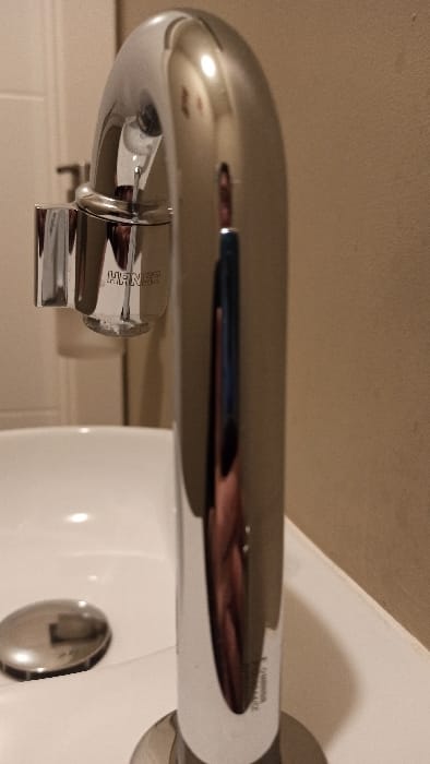 The image presents a close-up view of a modern, chrome-finished Hansa brand faucet installed over a white ceramic basin. The faucet exhibits a tall, arching design, with a small chrome cylindrical attachment protruding from its left side and a visible sensor-like element below the spout. A singular drop of water is suspended at the faucet's tip. A circular chrome drain cover is positioned in the lower-left area of the basin. The immediate background is a plain, light beige wall. The highly polished surface of the faucet prominently reflects a person's upper torso and head, seemingly wearing dark clothing, on its main vertical shaft, along with a distinct reflection of an overhead light source near the top curve. This scene is located in Florenville, Belgium.