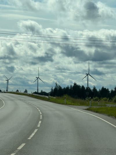 This media file depicts a landscape scene featuring a winding road, wind turbines, and power lines under a cloudy sky. The road, paved with asphalt, curves to the right and is marked with white dashed lines. The surrounding environment includes a grassy verge and a dense line of trees. Several wind turbines are visible in the distance, silhouetted against the sky. The sky itself is filled with a mix of light and dark clouds, suggesting a dynamic weather condition, possibly overcast or with intermittent sunlight. Power lines are also prominent in the upper left portion of the image, running parallel to each other. The lighting indicates it is daytime, though the cloud cover likely diffuses the sunlight. The scene conveys a sense of rural or open countryside, common in areas with renewable energy infrastructure. There is no visible text in the image.
