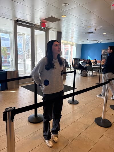 The image, rotated 90 degrees clockwise, depicts an indoor scene, likely a cafe or cafeteria, in Waltham, United States. In the foreground, a woman stands in a queue defined by black stanchions with silver tops. She wears a light grey sweater adorned with blue polka dots, dark pants, and light-colored sneakers, and looks forward or slightly upward with a neutral expression. A letter "Z" is visible on her sweater.
Behind her, another person stands in line. Further back, several people are seated at tables, some appearing to be using laptops or eating. The floor is tiled in a light beige pattern, and the ceiling features recessed lighting. Large windows along the left side of the frame reveal an outdoor view of another building and some trees, indicating it is daytime.
Notable details include a red "EXIT" sign mounted on the wall near the windows. A standing sign or menu board in the mid-ground prominently displays the word "ENTER," with other partially legible text like "YOU WANT" and "TEL." Inverted text visible through the window reads "CAFÉ DEROULLE." The setting suggests a casual, public environment where people are either waiting in line or dining/working.