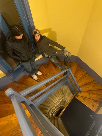 The image displays two individuals on a wooden spiral staircase within a building in Roanne, France. The walls are painted a bright yellow, contrasting with the muted blue-grey banister and railings of the staircase. The steps are crafted from light brown wood. On the left, an individual with long dark hair, dressed in a black puffer jacket, blue jeans, and white shoes, stands on a landing with hands in pockets, looking downwards. On the right, a second individual with long dark hair, wearing a black and white polka dot baseball cap, a dark grey buttoned blazer over a light grey shirt, faded blue jeans, and dark brown boots, is positioned on the staircase. This individual's left hand rests on the blue-grey railing, their left leg is bent with the foot on a lower step, and their right foot is on an upper step, while they look directly towards the camera. The staircase descends below the visible landing, revealing a tight spiral design with additional wooden steps and an inner wooden handrail. A blue-painted door frame or window frame is visible on the left side of the scene.