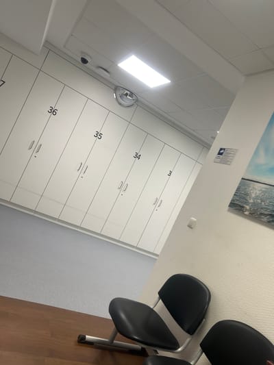 The image displays an unoccupied indoor room, likely a waiting area or changing facility, captured from a tilted perspective. The main focus is on a long wall lined with white, two-tier lockers, each bearing a dark numerical label; numbers 33, 34, 35, and 36 are clearly visible.
To the right, a row of black, upholstered chairs with silver metal frames is partially seen, suggesting a waiting or seating area. The floor transitions from a light grey, speckled surface near the lockers to a warm brown wooden floor under the chairs. The ceiling consists of white acoustic tiles, featuring a bright, rectangular fluorescent light fixture, a dome surveillance camera, and a circular convex security mirror.
Above the lockers, the wall is covered in textured white wallpaper. A landscape picture depicting a body of water under a sky is mounted high on this wall. Below the picture, a small rectangular sign features a blue and white icon, possibly indicating accessibility (resembling a person in a wheelchair), along with some illegible German text. A white wall switch or control panel is also visible on this wall. The overall impression is a clean, functional, and well-lit indoor space, typical of a public facility in Herne, Germany. No people or activities are present.