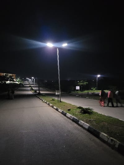 The image depicts a nighttime scene on a paved road in Gaba, Nigeria, illuminated by streetlights. A prominent, tall, grey streetlight pole with two bright luminaires stands centrally in the foreground, emanating strong artificial light. Attached to this pole is a small white sign displaying the number "20". The asphalt road extends into the distance, bordered on the right by a low concrete curb with alternating light and dark segments, separating it from a narrow strip of grass and sparse vegetation. Another streetlight with a single luminaire is visible further down the road on the right, next to another illegible white sign. On the far right side of the road, three indistinct figures are walking away from the camera, blurred by motion; one appears to be wearing reddish attire. On the far left, near the edge of the frame, two additional indistinct figures are partially visible next to a building. Multiple streetlights line the road as it recedes, suggesting a well-lit thoroughfare. The sky is dark, indicating a clear night.