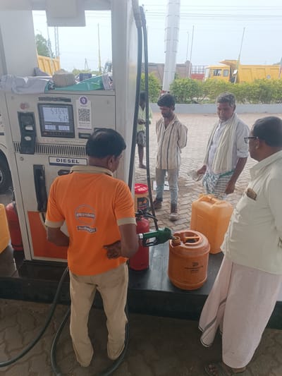 The image depicts a fuel station in Sugalimetta, India. A gas pump displays "DIESEL" and shows a transaction amount of 595.44 and a volume of 809.9 Ltr. A fuel attendant in an orange Indian Oil t-shirt is standing by the pump, with his hand on his hip. Two customers, dressed in traditional Indian attire, are also present. One man, wearing a striped shirt and light-colored pants, stands near the pump. Another man, wearing a white shirt and a white cloth draped around his neck, is holding a white cloth in his hand. Nearby, there are several large plastic containers, one of which is being filled with diesel from the pump. In the background, yellow trucks and greenery are visible. The scene appears to be taking place outdoors under an overcast sky.