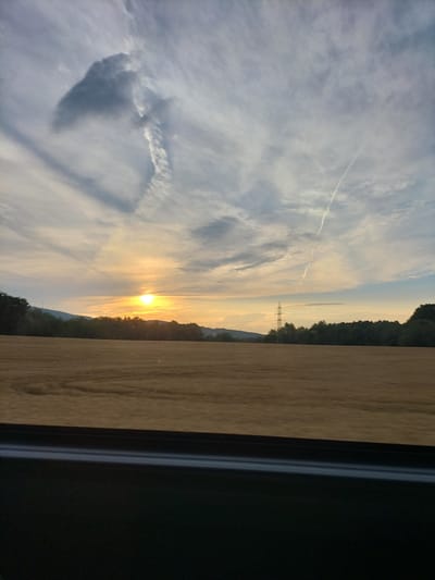 An eye-level, medium shot captures a rural landscape at sunset. The sun, a bright orb, is positioned above the horizon, casting a warm glow across a vast field of harvested grain, its texture appearing textured and golden-brown. A dark line of trees and rolling hills forms a backdrop, with a distant electrical pylon visible amidst the foliage. The sky is a dramatic canvas of scattered clouds, some wispy and white, others darker and more textured, interspersed with two prominent contrails from aircraft, one sharply defined and the other more diffused. The lower portion of the frame is dominated by the dark, indistinct interior of a vehicle, likely a car, suggesting the photograph was taken from a window. The overall atmosphere is tranquil and expansive, indicative of a scene in the countryside, potentially near Horní Třebonín, Czechia.