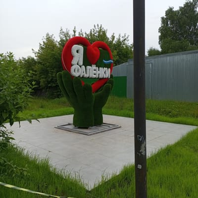 The image shows a monument in the city of Falenki, Russia, dedicated to the city itself. The monument features a large, stylized red heart atop two green, hand-like sculptures made of artificial turf. Inside the heart, the white Russian letters "Я" and "ФАЛЁНКИ" are prominently displayed, with "Я" meaning "I" and "ФАЛЁНКИ" being the name of the city. A small blue and white wavy line is positioned between the two words. The monument is situated on a square paved area, surrounded by green grass and trees. In the background, there is a grey corrugated metal fence and more trees. A dark, weathered metal pole with some graffiti is visible in the foreground, partially obscuring the view of the monument. The sky is overcast, suggesting a cloudy day. The overall scene depicts a civic monument celebrating local identity in the urban environment of Falenki.