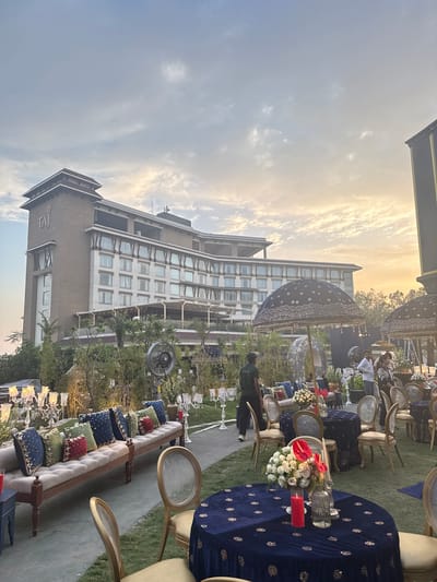 An outdoor event is captured during the golden hour, likely on a hotel lawn or garden. The scene is set for a formal gathering, possibly a reception or party, with numerous decorated tables and seating arrangements.

The central subjects include several round and rectangular tables draped in rich dark blue tablecloths, many featuring intricate gold patterns. Elegant gold-framed chairs are placed around these tables. Floral centerpieces, composed of white roses and striking red anthurium flowers, adorn the tables, accompanied by tall red candles and clear glass candelabras or vases. Long, tufted benches are also present, lavishly decorated with an array of plush cushions in shades of blue, green, and red, all embellished with gold embroidery. Large, decorative dark blue umbrellas or canopies, patterned with white designs, provide a festive touch and potential shade.

Several individuals are visible throughout the space; some are seated at tables, while others stand, suggesting both attendees and possibly event staff. One person in a dark green outfit is clearly visible in the foreground.

In the background, a modern, multi-story hotel building dominates the left side of the frame, its numerous windows reflecting the soft light. The sky above is a blend of pale blue and warm golden hues, indicative of late afternoon or early evening. The overall atmosphere appears elegant and celebratory.

A prominent detail is the text "TAJ" visible on the side of the hotel building, identifying the venue as part of the Taj Hotels chain, consistent with the provided location context of Dehradun, India.