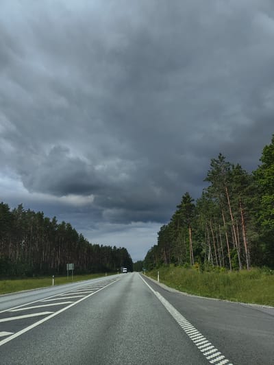The image shows a two-lane asphalt road stretching into the distance, bordered by a dense forest of pine trees on both sides. The sky is heavily overcast with dark, dramatic storm clouds, suggesting an impending rain shower. White road markings, including dashed lines and a solid white line, delineate the lanes. On the left side of the road, there are crosswalk markings. A white van is visible in the distance on the right lane. The overall environment appears to be rural, consistent with the outskirts of Uulu, Estonia, a region known for its natural landscapes and forests. The scene captures a moment of atmospheric tension before a storm, with the road serving as the central element connecting the viewer to the dense, dark forest under the dramatic sky.