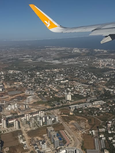 The image is an aerial view, taken from an airplane, looking down at an urban and semi-urban landscape. The most prominent object in the foreground is the wing of the aircraft, painted with a yellow and white design featuring a white Pegasus-like creature. The wing extends diagonally from the lower left to the upper left of the frame.

The scene below is a sprawling development, appearing to be a mix of residential areas and some commercial or industrial buildings. There are numerous white or light-colored buildings with dark roofs, densely packed in some areas and more spread out in others. Roads and pathways are visible, winding through the urban fabric. Some areas show construction sites with foundations and unfinished structures. Green patches, possibly parks or agricultural land, are interspersed within the developed areas. The overall impression is of a sunny day with clear skies, suggested by the bright lighting and the deep blue of the sky. The image is oriented vertically, with the wing at the top left and the cityscape extending downwards and to the right. The location is identified as Muratpaşa, Turkey, which implies a coastal or near-coastal region based on typical Turkish geography. No people are discernible in the image, and no text is visible.