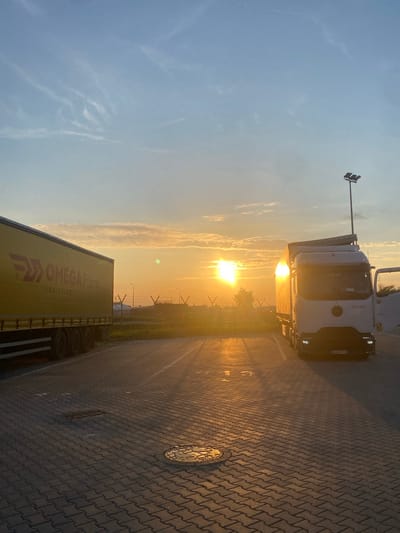 The image depicts a scene at sunrise in the city of Niepołomice, Poland. The sky is a soft blue with wispy clouds and the sun is low on the horizon, casting a warm, golden light. In the foreground, a paved area, likely a parking lot, is visible with interlocking brick pavers. Two trucks are parked on this lot. The truck on the left is a large, yellow semi-trailer with the "OMEGA Pilzno LOGISTICS" logo prominently displayed. The truck on the right is a white semi-truck with its driver's side door open. The sun's rays create long shadows across the paved surface. In the background, there are signs of industrial or logistical infrastructure, including fencing and lighting poles. The overall impression is one of the start of a workday in an industrial area, bathed in the gentle light of dawn.