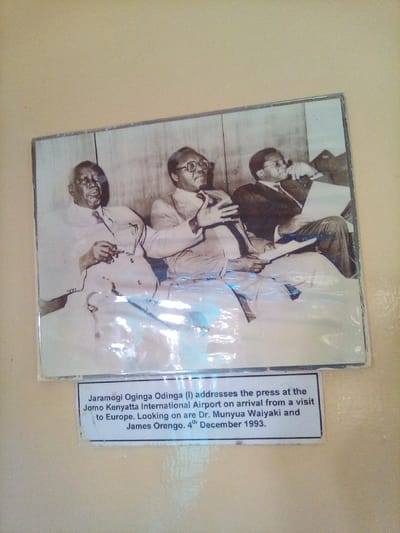 A rectangular, horizontally oriented photograph, appearing to be a black-and-white or sepia print, is mounted on a plain, light-colored wall, possibly beige. The photograph is covered by a reflective, transparent material, causing glare across its surface. Below the photograph, a white caption card with black text is attached.
The photograph depicts three adult males seated side-by-side, appearing to be in an indoor setting with a paneled background. From left to right:
1. **Jaramogi Oginga Odinga:** An older man, wearing a light-colored suit, collared shirt, and tie. He looks towards the viewer's left with his mouth slightly open, holding a pair of glasses in his left hand.
2. **Dr. Munyua Waiyaki:** A man wearing a suit, collared shirt, and glasses. He looks towards the right, with his right hand raised and open, gesturing. Papers are visible in his lap.
3. **James Orengo:** A man wearing a dark suit, collared shirt, and glasses. He looks down at papers held in his lap with his left hand, while his right hand holds a pipe near his chin.
The caption beneath the photograph reads: "Jaramogi Oginga Odinga (l) addresses the press at the Jomo Kenyatta International Airport on arrival from a visit to Europe. Looking on are Dr. Munyua Waiyaki and James Orengo. 4th December 1993."
This image, comprising the photograph and its caption, is located in South Sakwa ward, Kenya.