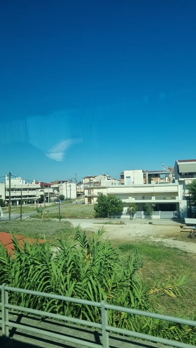 The image captures a view from a moving vehicle, likely a bus or car, looking towards a suburban area under a clear blue sky. In the foreground, lush green reeds and a metal railing suggest the perspective is from a roadway or bridge. Beyond this, a sparsely vegetated area gives way to a street lined with multi-story buildings. These buildings appear to be residential or commercial, with a mix of white and cream facades, balconies, and some with awnings. Power lines and poles are visible, indicating typical urban infrastructure. Further in the background, more buildings are visible, some with red tiled roofs. A light blue vehicle is parked on the street, and a green trash bin is also present. The overall scene depicts a sunny day in a residential district of Larissa, Greece, with the characteristic architecture and infrastructure of the region.
