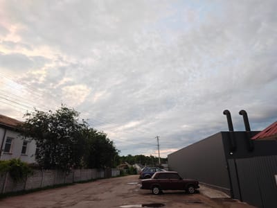 The image depicts an outdoor scene in the city of Rzhyshchiv, Ukraine, likely in an industrial or commercial area, possibly near a parking lot. The sky is filled with a textured pattern of light gray and white clouds, with hints of pale pink and yellow suggesting either dawn or dusk. In the foreground, a dirty, uneven asphalt surface is visible, with several puddles of water reflecting the sky. A dark red, four-door sedan is parked on the asphalt. To the right, a long, dark gray building with a modern, angular design dominates the mid-ground. Two large, black, flexible exhaust pipes extend vertically from the roof of this building. A section of dark gray corrugated metal fencing is attached to the building. Further to the left, a concrete fence with a decorative pattern runs along the edge of a property. Behind this fence, a two-story white building with two windows is partially visible, partially obscured by dense, dark green foliage. Utility lines and poles are visible stretching across the sky in the background, and a forest or cluster of trees forms a distant backdrop. Another smaller blue car is visible parked further down the dirt road.