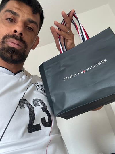 A male individual, appearing to be in his 30s or 40s with dark hair and a greying beard, is depicted in a close-up, low-angle selfie or portrait in an interior setting in Oberasbach, Germany. He is wearing a white collared shirt, possibly a sports jersey, featuring the number "23" in black on the left side and a dark emblem on the right shoulder. His right hand, adorned with a silver ring on the ring finger, holds a dark grey Tommy Hilfiger shopping bag. The bag's handles are striped with red, white, and dark blue. The "TOMMY HILFIGER" logo, featuring the brand's iconic flag, is prominently displayed in white text on the bag's surface. The background consists of a plain, light-colored wall. The individual maintains direct eye contact with the camera.