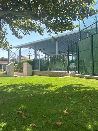 The image depicts an outdoor sports facility, likely a tennis or padel court, under a large, arched roof structure. The facility is situated in a well-maintained grassy area, with several fallen leaves scattered on the lawn. The foreground is dominated by a lush green lawn bathed in sunlight, with the dappled shadows of tree leaves cast upon it. In the middle ground, there are two prominent, angular concrete pillars, serving as an entrance or pathway marker. Beyond these pillars, the sports court area is visible, enclosed by green fencing and netting. A dense green hedge runs along the perimeter of the court. The background reveals the roof structure supported by metal beams and columns, with more netting and fencing visible underneath. The sky is clear and blue. While the specific location of Villa del Prado, Spain, is mentioned, no visual elements in the image directly confirm this geographical context beyond a general European aesthetic. There are no people or animals visible in the scene, and no actions or interactions are taking place.