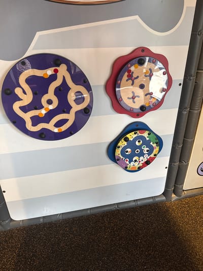 The image shows a play area with interactive wall games. There are three circular games mounted on a white and blue striped wall. The top left game is red and has a clear plastic covering small wooden shapes. The top right game is blue and has a cartoonish food theme. The bottom game is purple and has a wooden track with small balls. The play area is located in Mountain Park, United States of America.