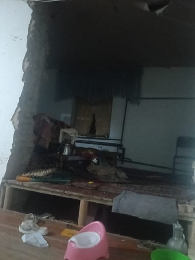 The image depicts a damaged interior space, possibly a room, with a large section of the wall or ceiling collapsed. The exposed interior structure suggests recent destruction or neglect. Debris is visible within the room, including fabric items and what appears to be a metal kettle or pot. A shelf-like structure is present, holding various objects, some of which are indistinct. In the foreground, a pink potty chair, a set of keys on a white object, and a small paper item are visible on a wooden surface. A glass bottle or jug with a lid is also present. The overall impression is one of disarray and damage. There is no clear indication of the city of Altinkul, Uzbekistan, within the visual content of the image itself.