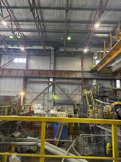 The image shows the interior of a large industrial facility with a corrugated metal roof and walls. The space is filled with various machinery, pipes, and metal structures, suggesting it is a processing or manufacturing plant. Yellow railings and stairs are prominent, indicating elevated walkways and platforms. There are also large ducts and ventilation systems visible. The lighting appears to be a mix of overhead industrial lights and some natural light filtering through windows. The overall impression is one of a busy, functional industrial environment. The location is identified as KOKENTAU RURAL DISTRICT, KAZAKHSTAN.