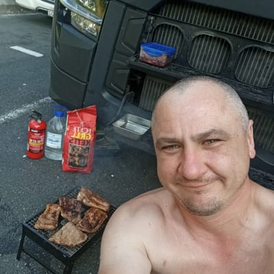 A man is taking a selfie in front of a large truck, with a small barbecue grill in the foreground. He is shirtless and has short hair and a mustache. On the grill are several pieces of cooked meat. Next to the grill are a bag of charcoal, a bottle of water, and a fire extinguisher. A blue plastic container with raw meat is placed on the truck's grille. The scene appears to be outdoors, possibly in a parking lot, given the asphalt ground and the presence of a truck. There is a faint hint of smoke in the air, suggesting the barbecue is in use or has recently been used. The lighting suggests it is daytime, likely late afternoon or early evening. The man has a slight smile on his face, appearing relaxed. Text visible on the bag of charcoal reads "ECILE GRILL" and "KETLEY 2KG". The fire extinguisher has "SOIDA" written on it.