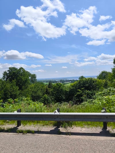 An elevated, wide shot shows a scenic view from a roadside pull-off overlooking a valley on a sunny day. In the foreground, a metal guardrail runs horizontally across the frame, casting shadows onto the asphalt road surface below. Behind the guardrail, lush green foliage, including trees and ferns, dominates the midground. Beyond the foliage, a vast expanse of water, likely a lake or river, stretches across the horizon, dotted with distant buildings and smaller bodies of water. The sky above is a vibrant blue, punctuated by fluffy white cumulus clouds. The scene suggests a peaceful, natural environment with signs of human habitation in the distance. The location is identified as Grenville-sur-la-Rouge, Canada.
